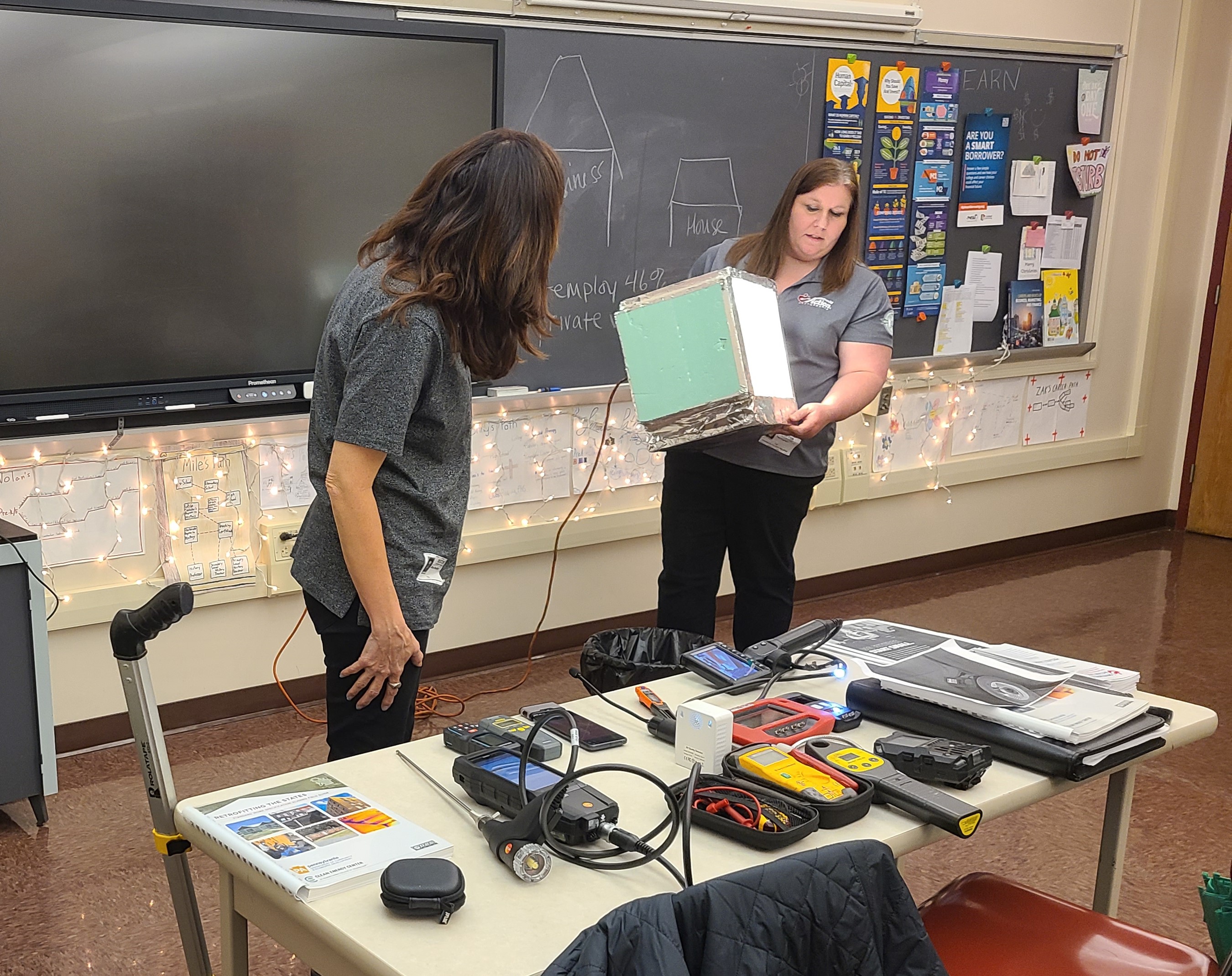 Tina Kephart, Weatherization Director, handles an insulation box while Diana McKillop, Quality Control Inspector, explains various insulation factors while demonstrating how infrared cameras display heat variations in rooms and are used to help determine where better insulation will reduce heat and energy loss.